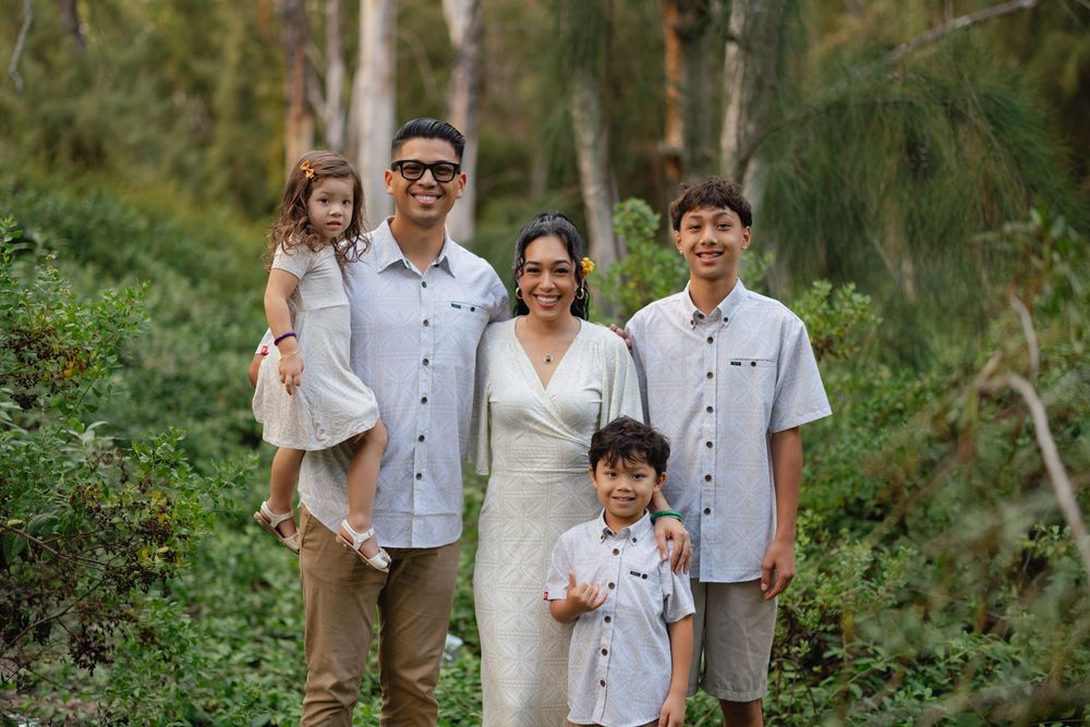 Family of five smiling outdoors in a lush green forest setting, wearing coordinated casual outfits for a portrait.