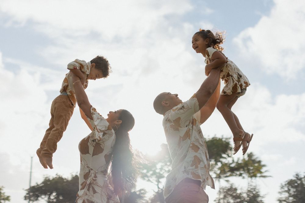 Two parents joyfully lift their children up in the air outdoors on a sunny day with clouds in the background.