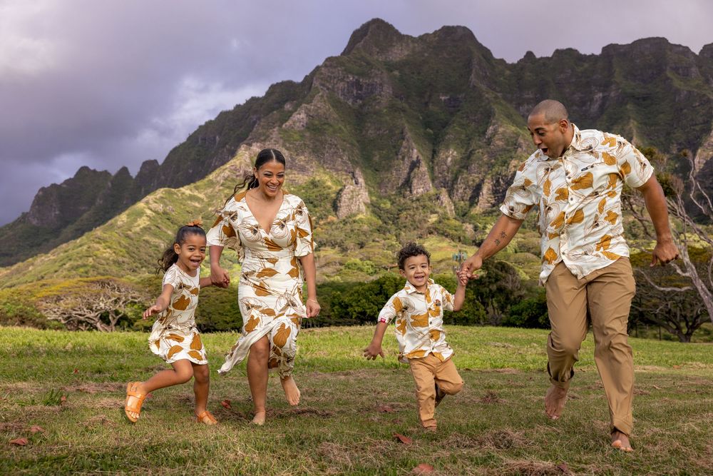 Family in matching tropical outfits joyfully running together on green lawn with dramatic mountain backdrop in Hawaii.