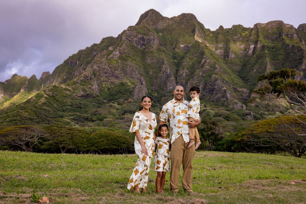 Family of four posing in matching floral outfits with lush green mountains backdrop in Hawaii.
