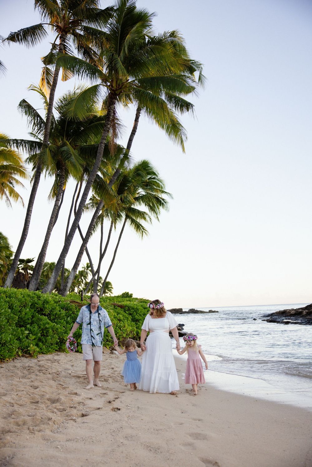 Family in wedding attire walks barefoot on a tropical Hawaiian beach at sunset with palm trees swaying in the breeze.