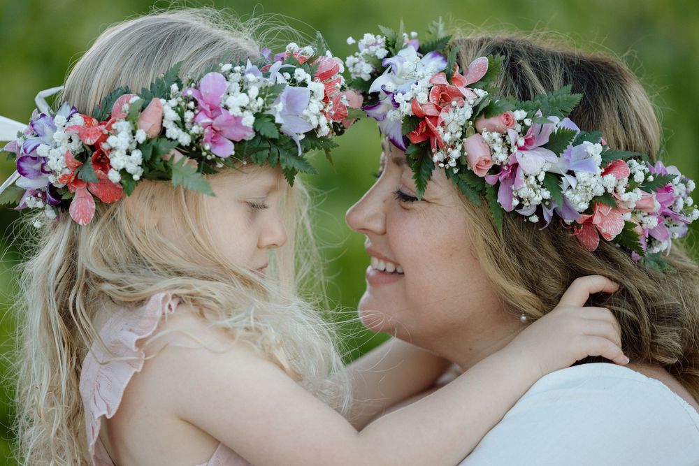 A mother and daughter wearing matching floral crowns share a tender moment outdoors.