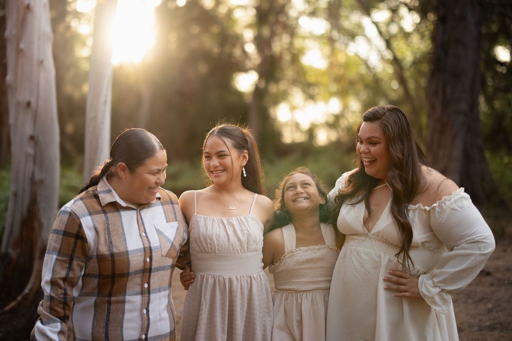 Four women laughing together outdoors in golden sunlight among trees, wearing casual and flowy dresses.