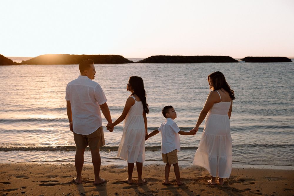 Family of four holding hands on a sandy beach at sunset, wearing white outfits, with calm ocean waters in the background.