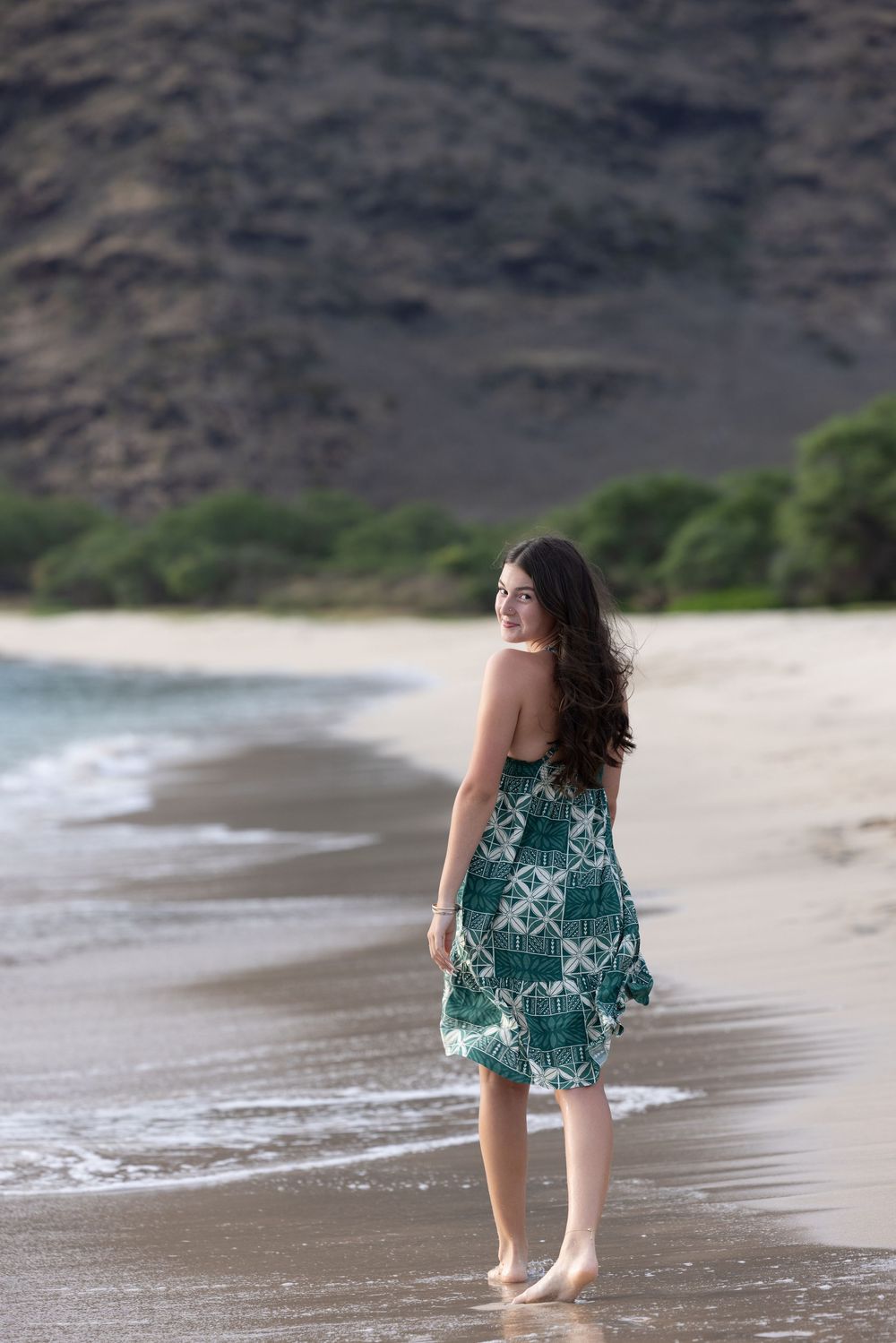 Young woman in green patterned sundress walking barefoot on sandy beach with mountain backdrop.