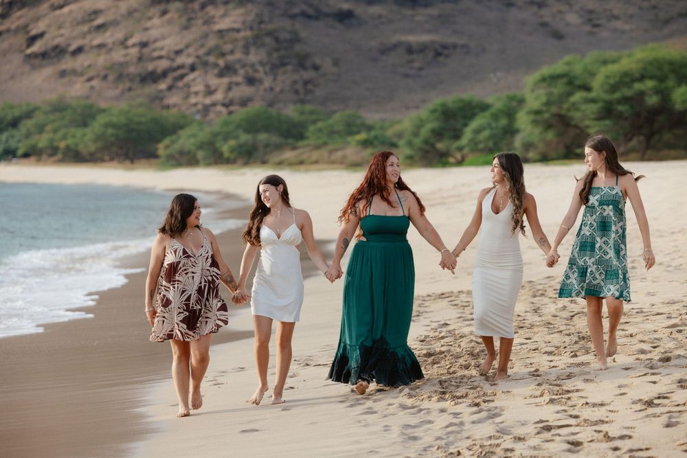 Group of five women in colorful dresses holding hands on a sunny Hawaiian beach with mountain backdrop.