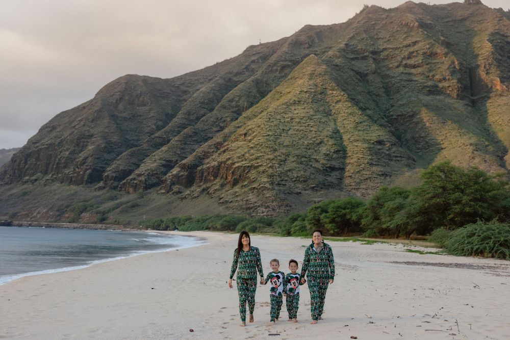 Family in matching pajamas walking on a sandy beach with lush green mountains in the background.