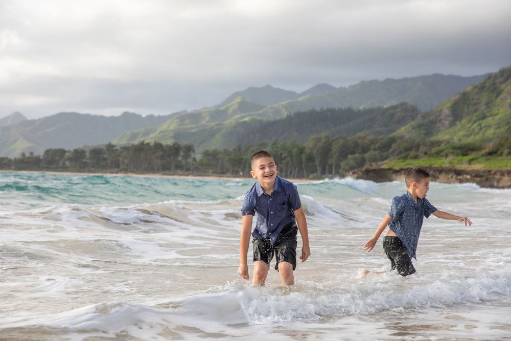 Two young boys laughing and playing in ocean waves on a Hawaiian beach with lush green mountains.
