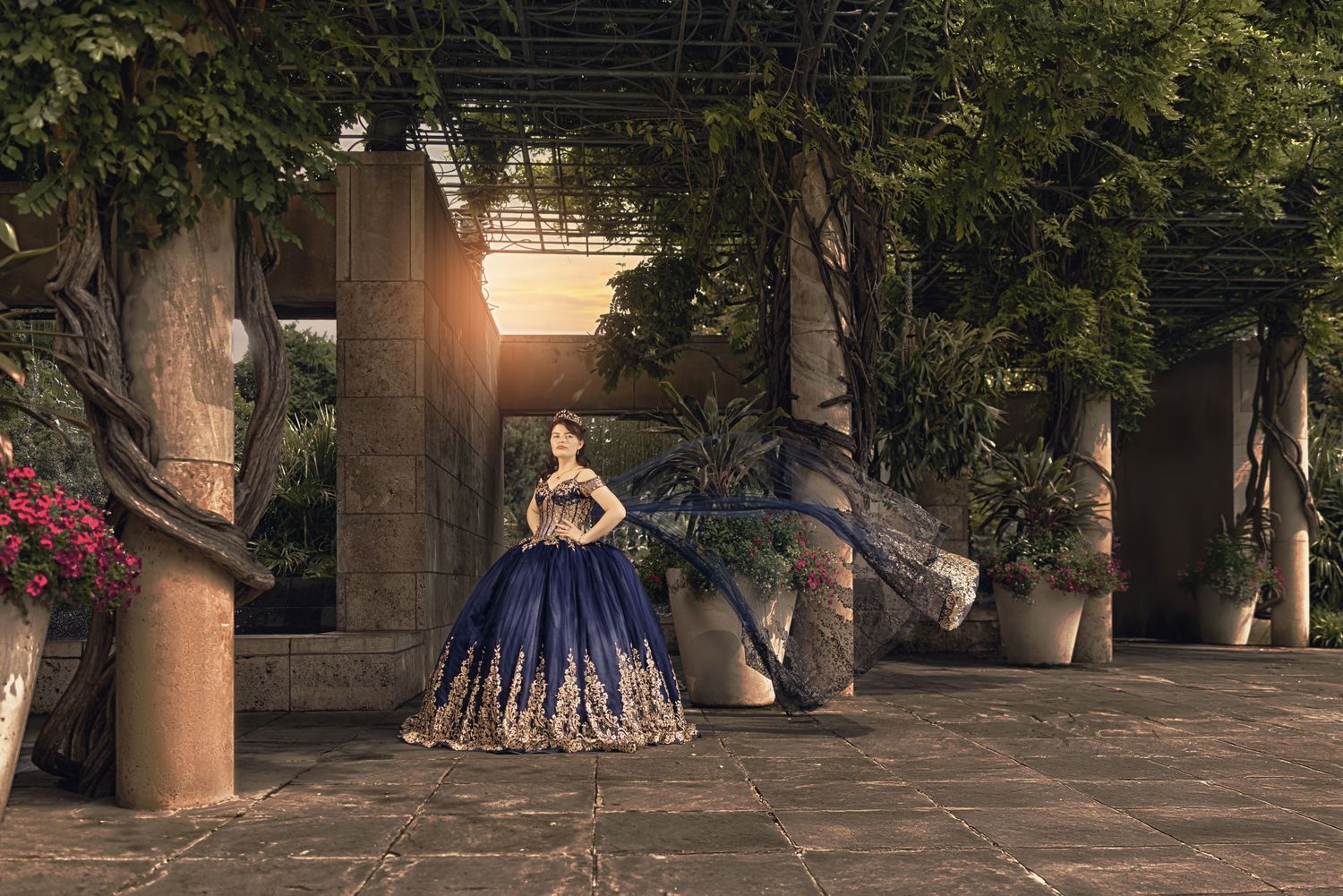 A teen in a navy blue and gold embroidered Quinceañera dress poses under a lush garden pergola with a flowing cape at golden hour.