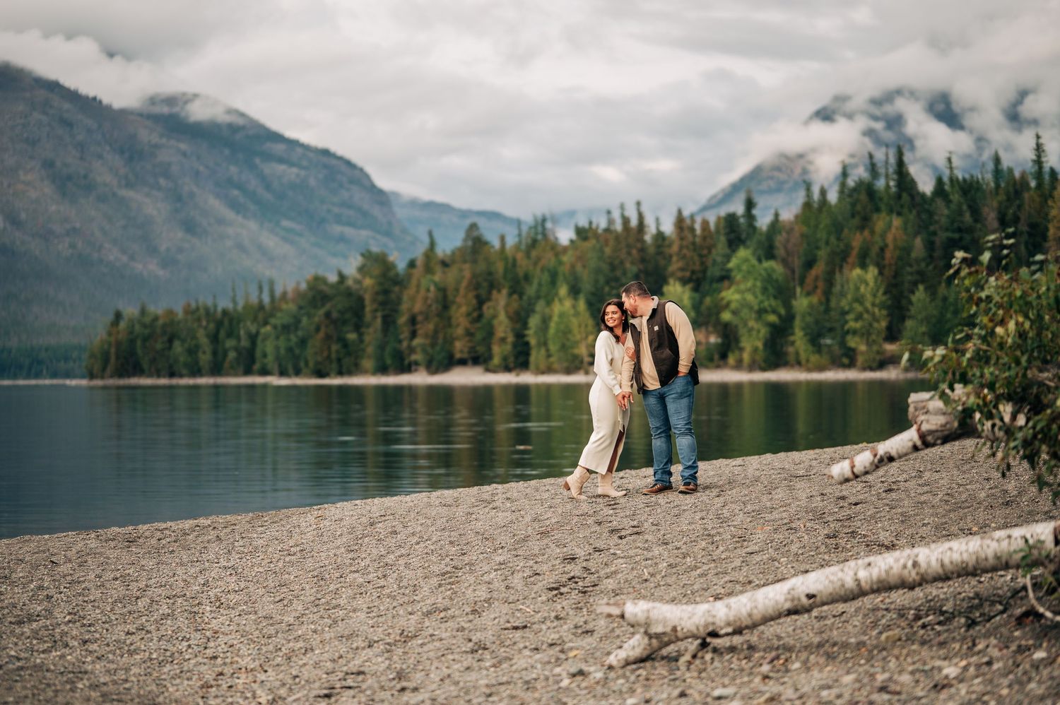 Couple embracing on a rocky lakeside beach with evergreen forest and misty mountains in the background.