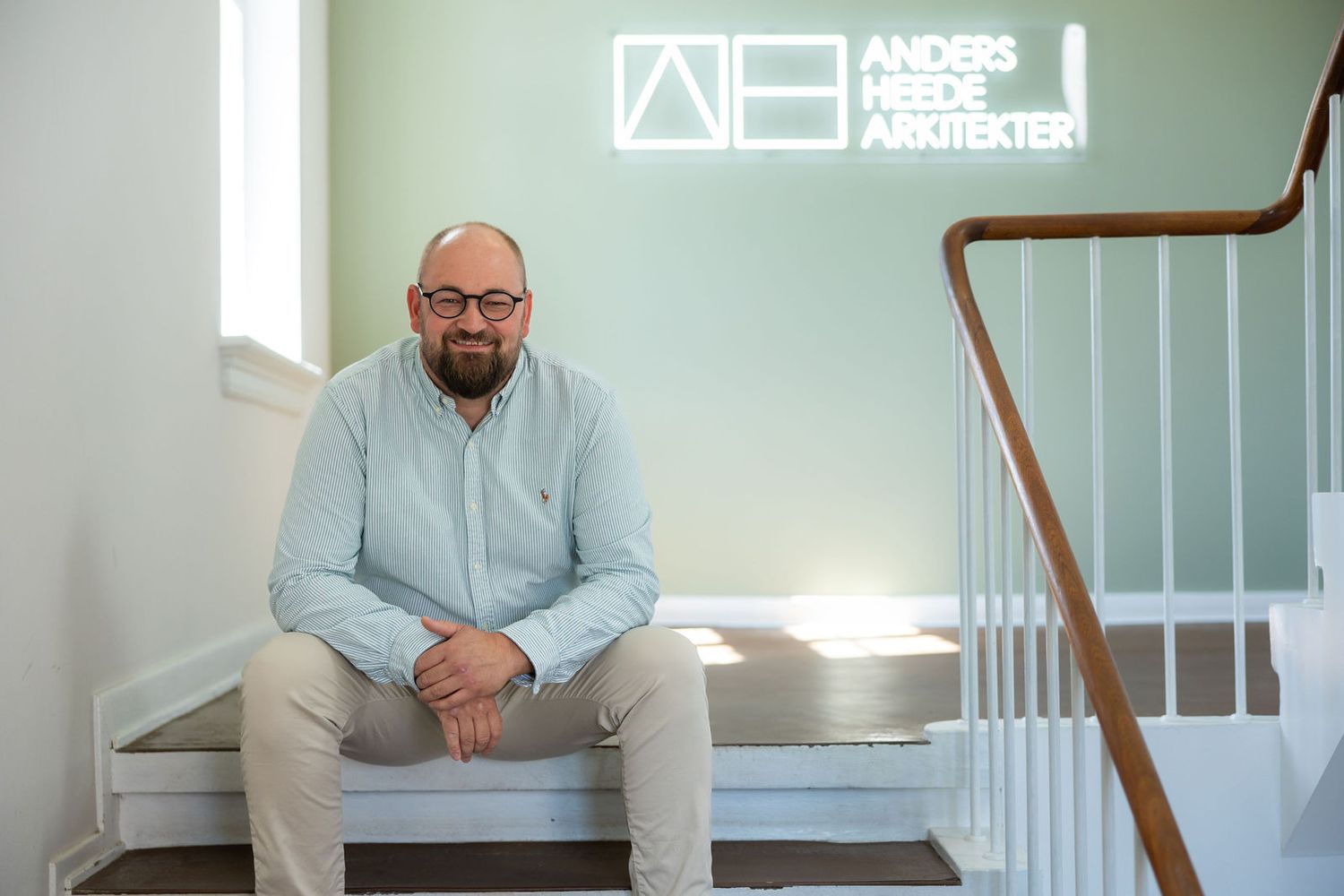 Man sitting on stairs beneath Anders Heede Arkitekter logo sign in a modern office interior.