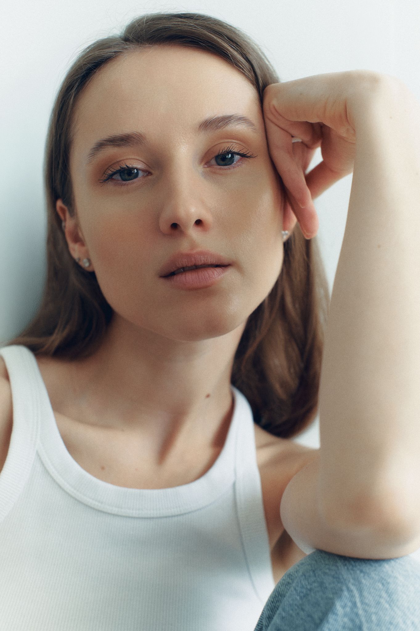 Young woman in white tank top resting hand against face, natural makeup, casual portrait against light background.