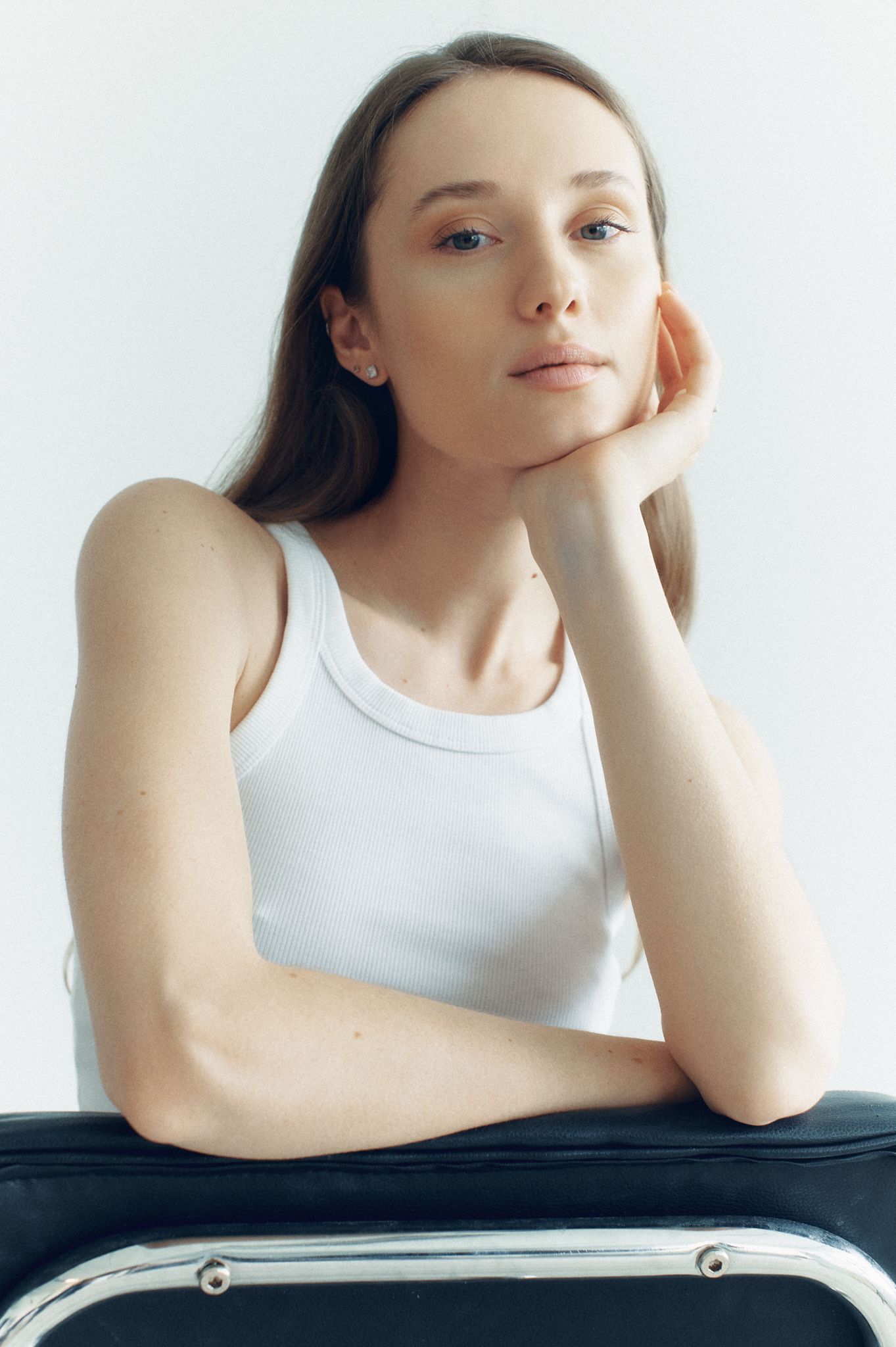 Young woman in white tank top resting chin on hand, sitting pensively against a light background.