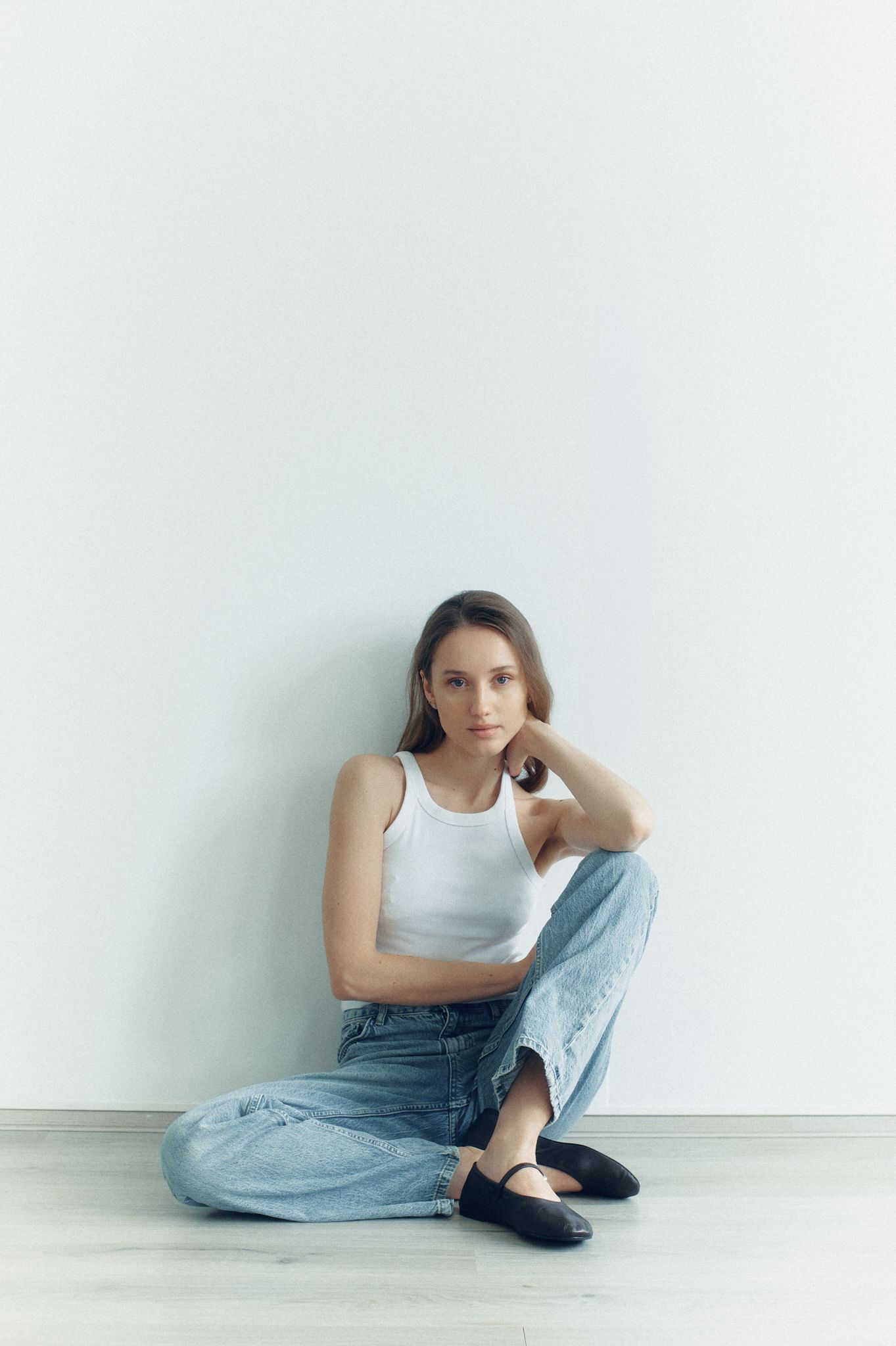 Young woman in white tank top and jeans sitting on floor against white wall, looking at camera.