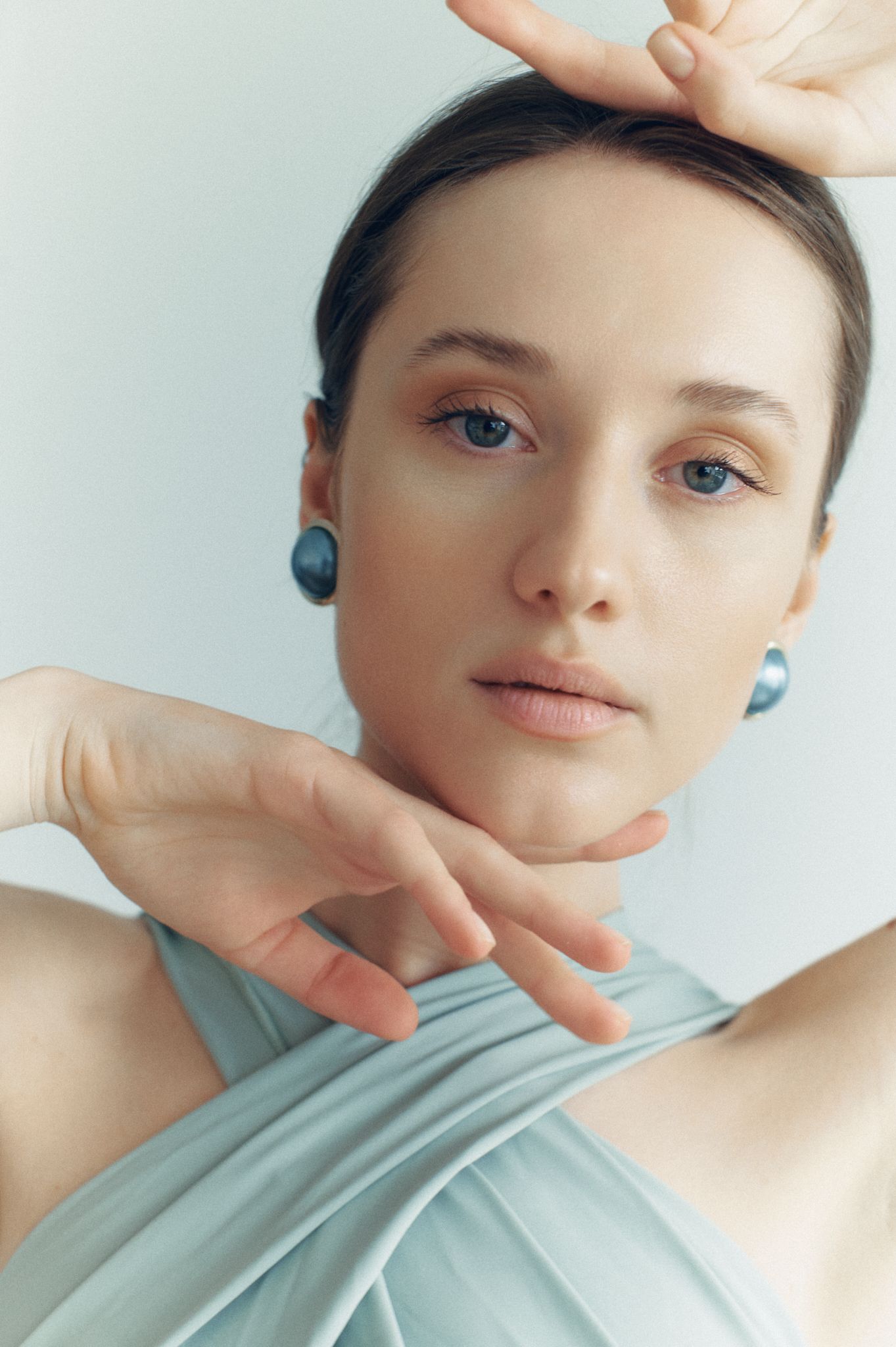 Young woman with blue earrings and minimal makeup poses gracefully in a light blue draped top against a soft background.