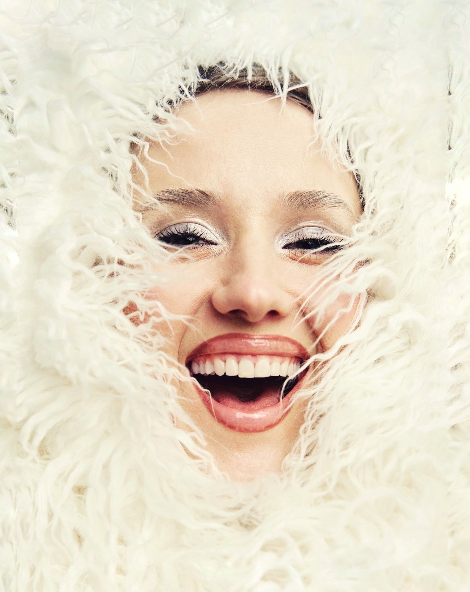 Woman laughing joyfully, surrounded by fluffy white feathers, wearing dramatic white eye makeup.