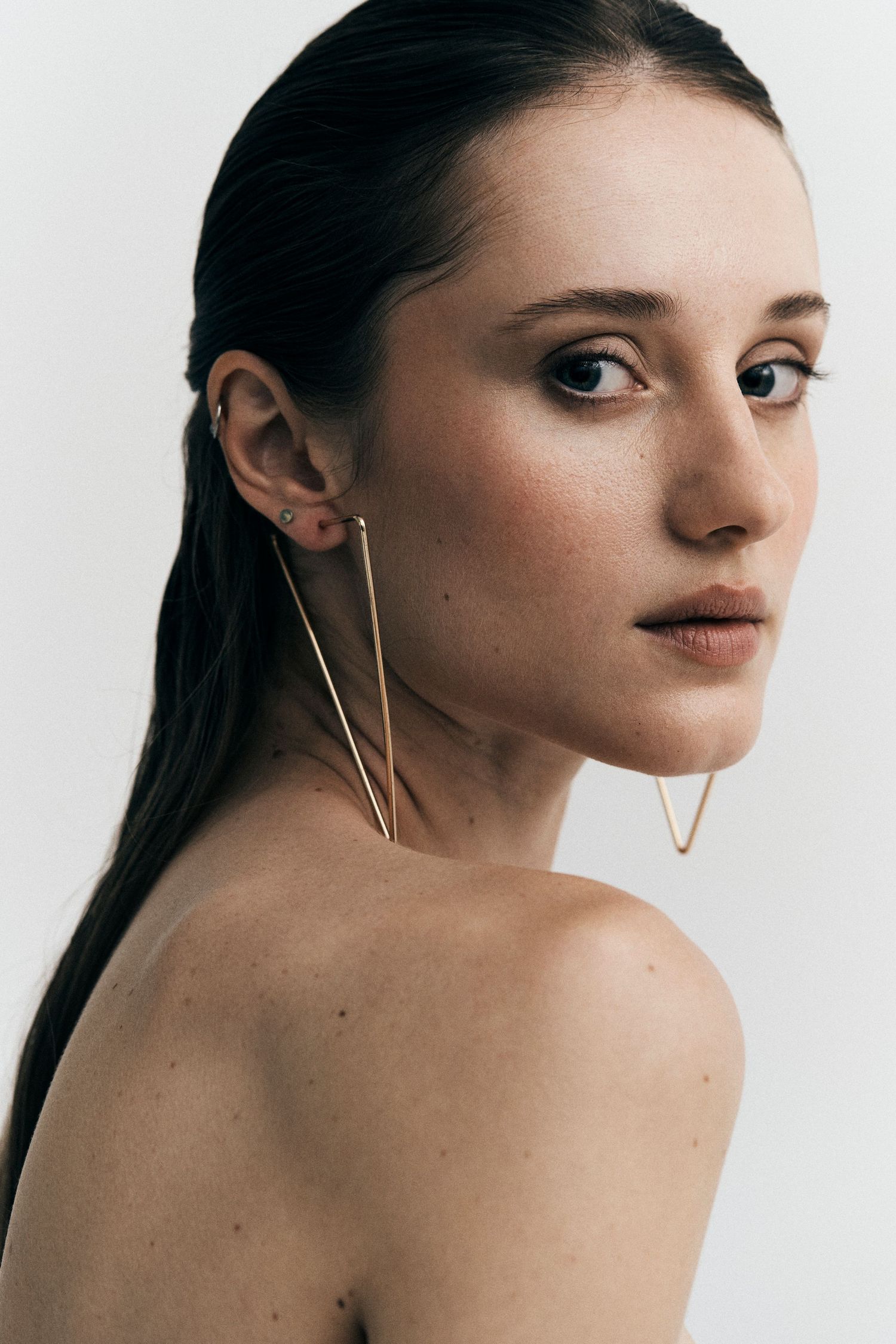Young woman wearing gold geometric triangle ear cuff earrings, minimal makeup, dark hair pulled back, light background.