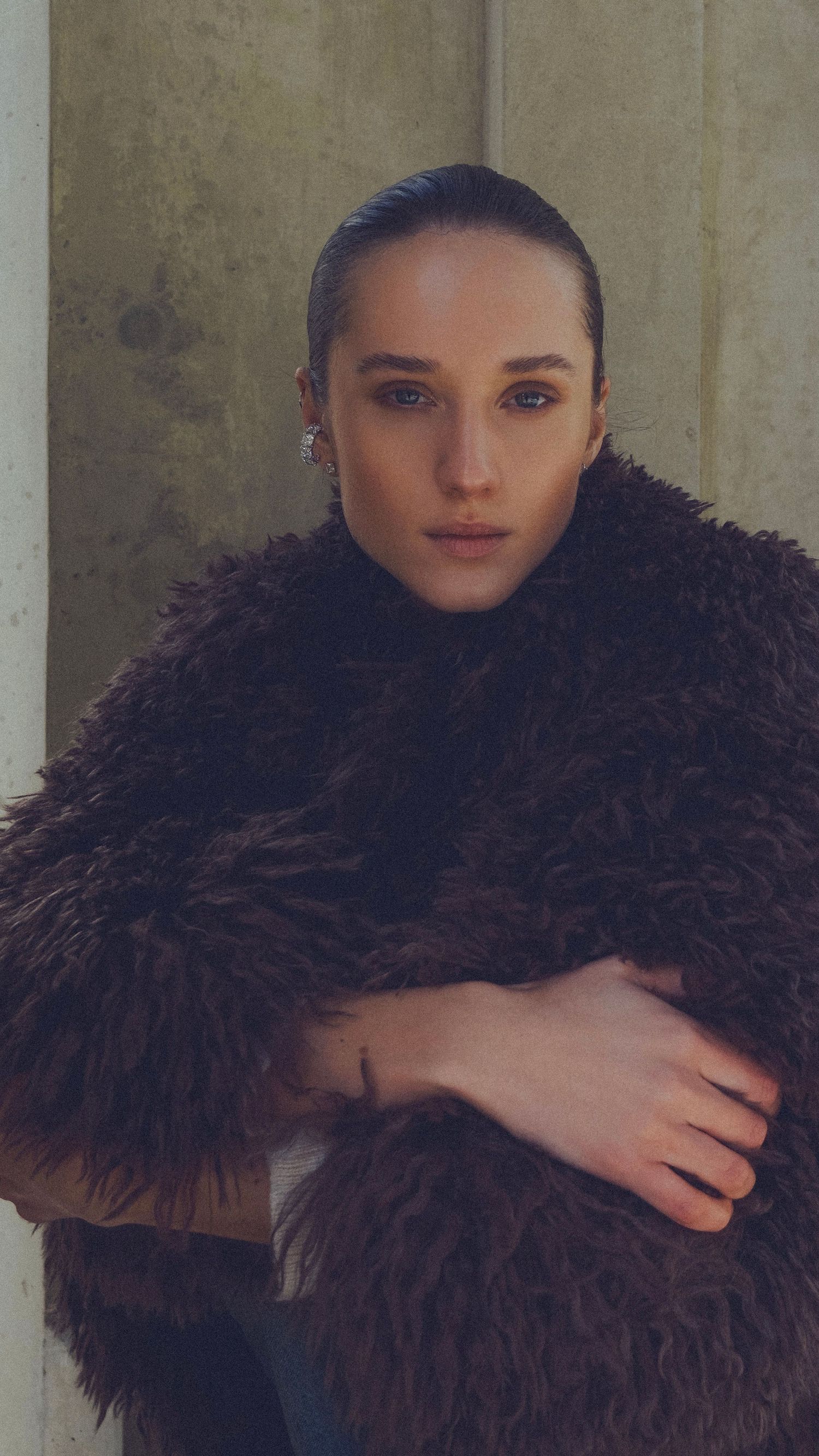 Young woman in dark fur coat with slicked-back hair poses confidently against a neutral wall.