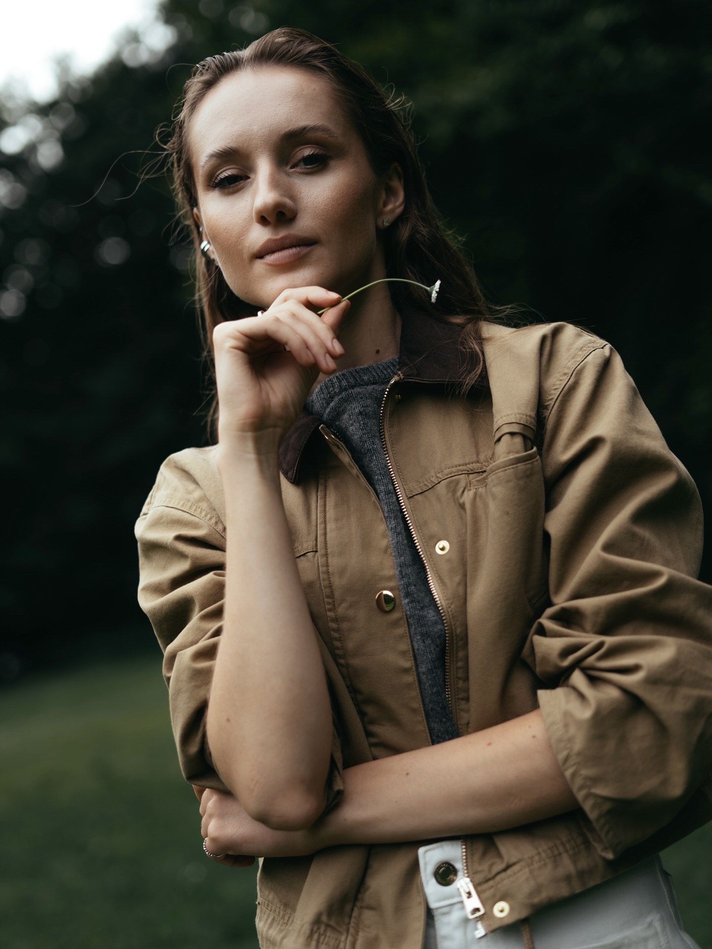 Young woman in tan jacket holding glasses outdoors, posing thoughtfully against a lush green background.