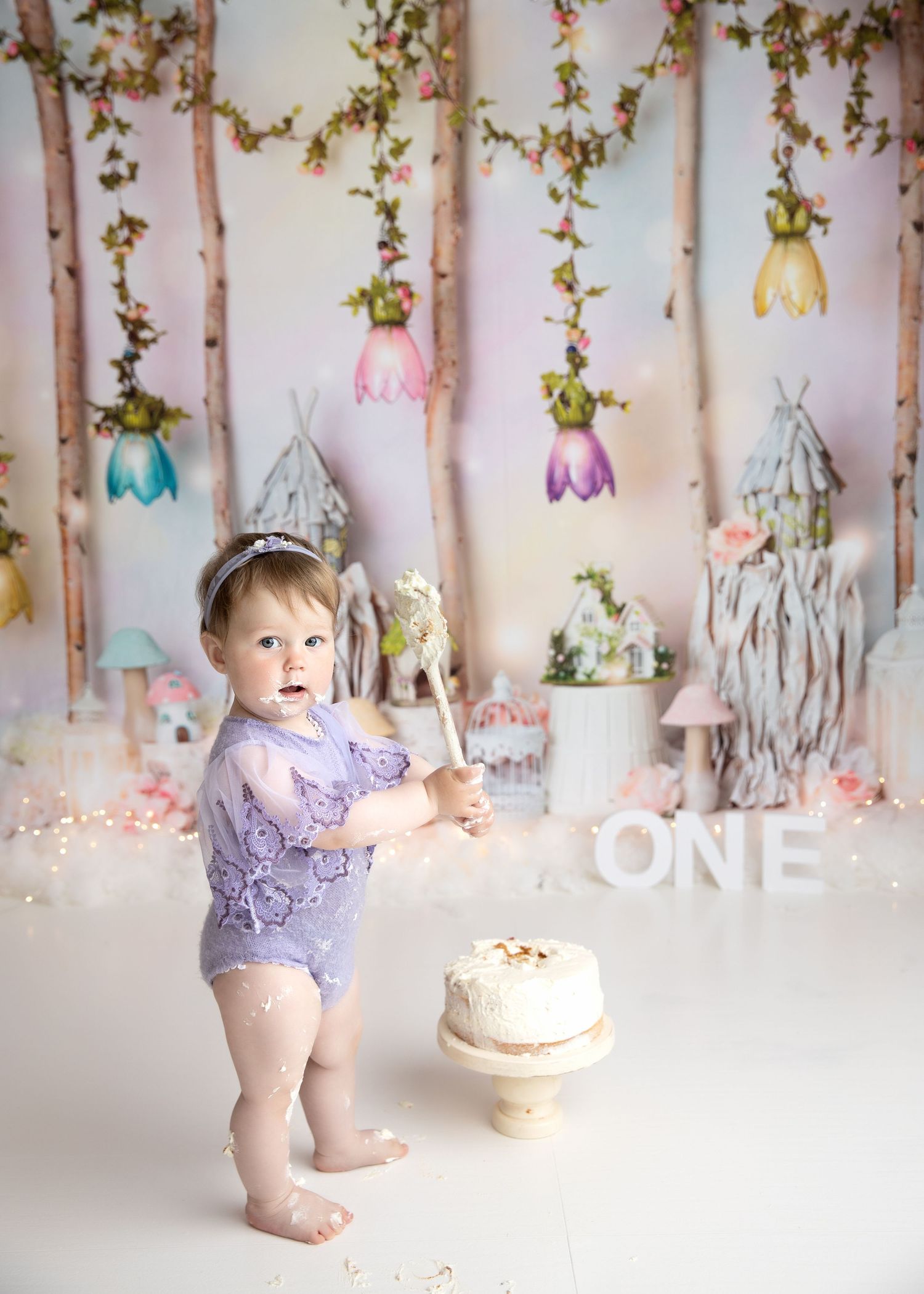 Baby girl in purple lace romper stands by first birthday cake with whimsical fairy tale backdrop and ONE sign. Cake Smash Photos Cork