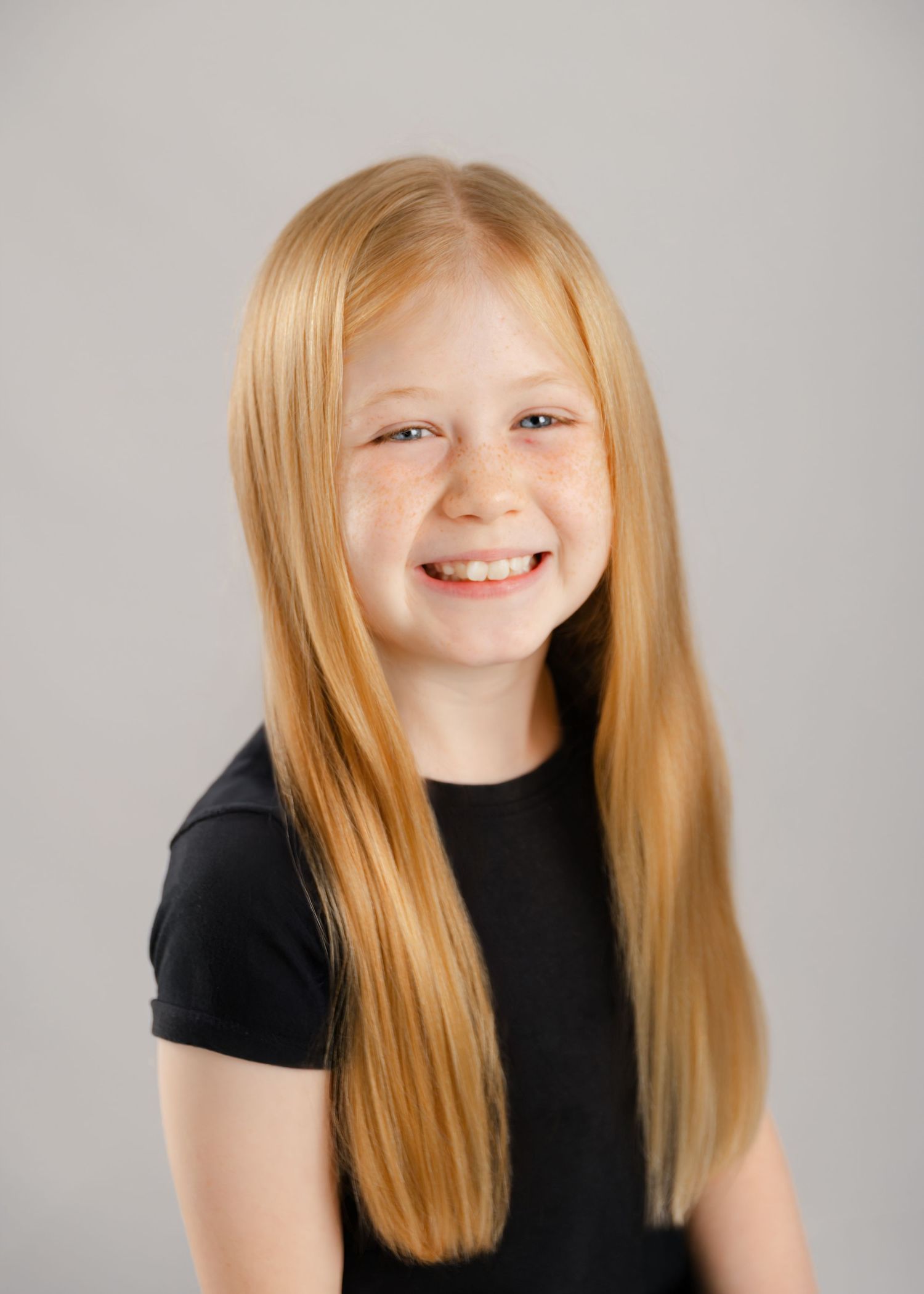 Smiling young girl with long straight blonde hair and freckles wearing a black top against a gray background.