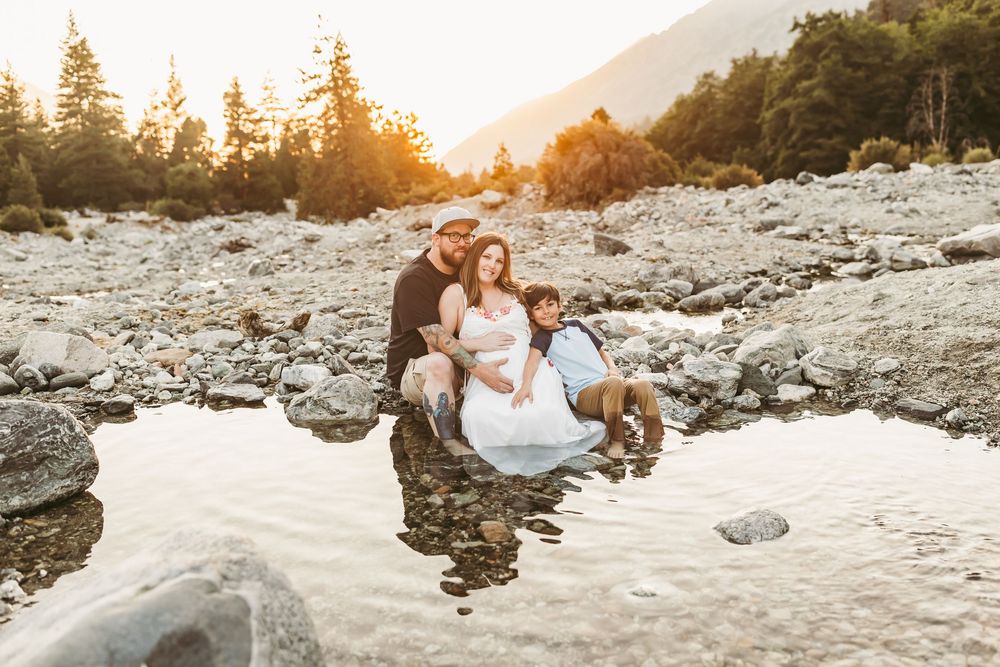 Family maternity photo session at golden hour by a mountain river, with warm sunset light filtering through pine trees near redlands ca