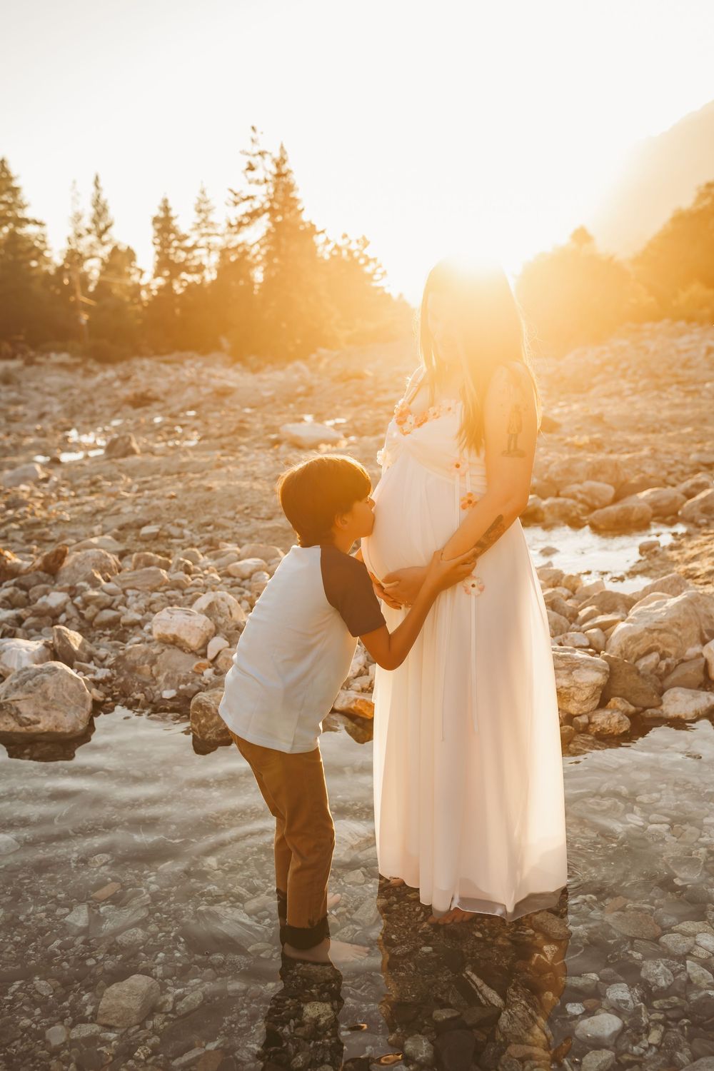 Boy kissing pregnant mothers belly outdoors in golden sunset light by a rocky mountain river in the inland empire