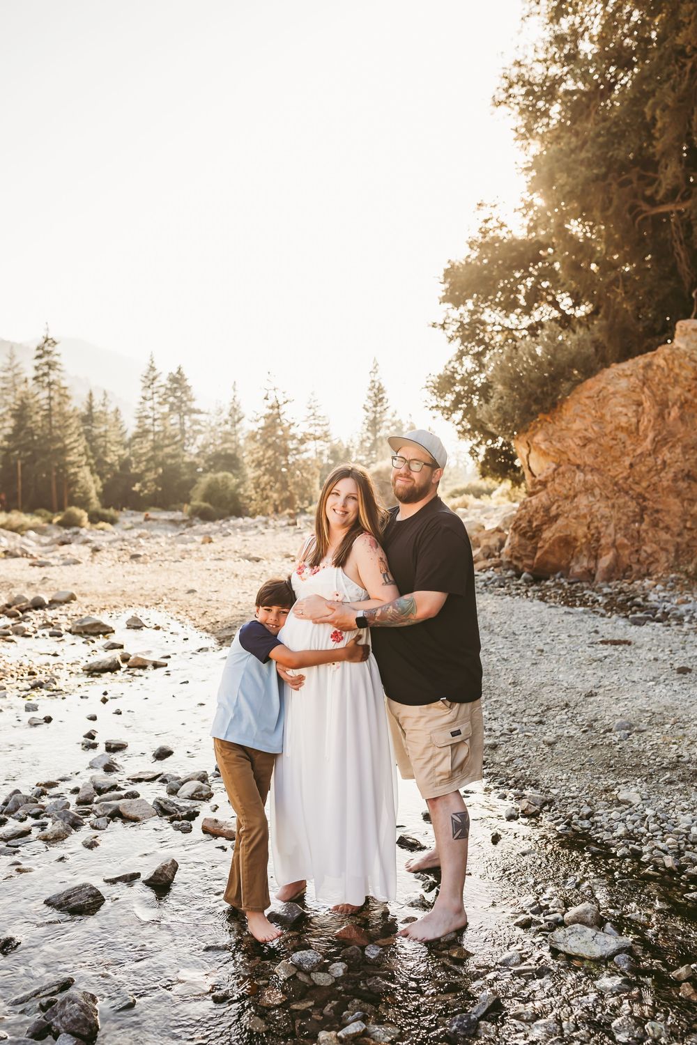 Family of three standing on rocky riverbed with golden sunset light filtering through pine trees with heidi grace photography