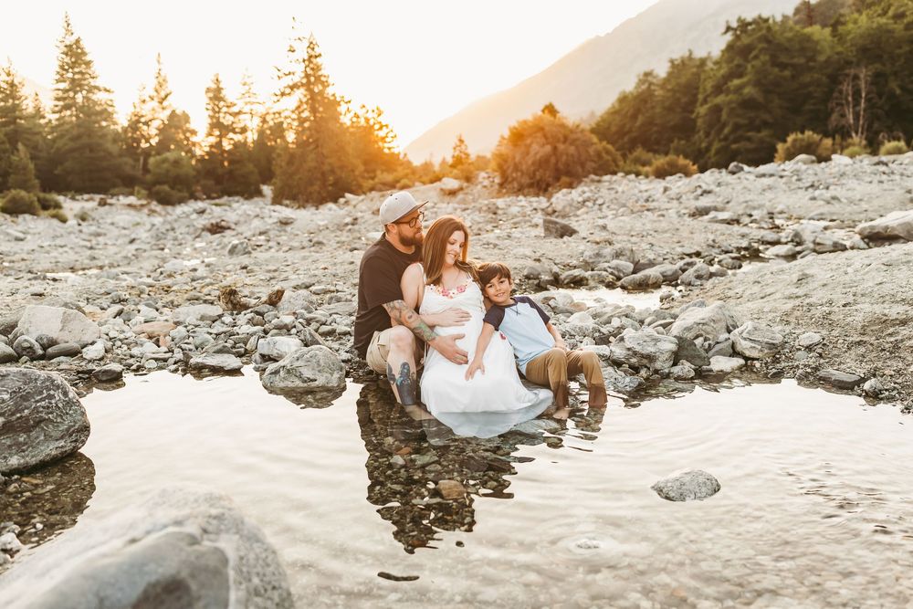 Family maternity photo shoot by a rocky mountain stream at golden hour sunset with warm light filtering through pine trees with heidi grace