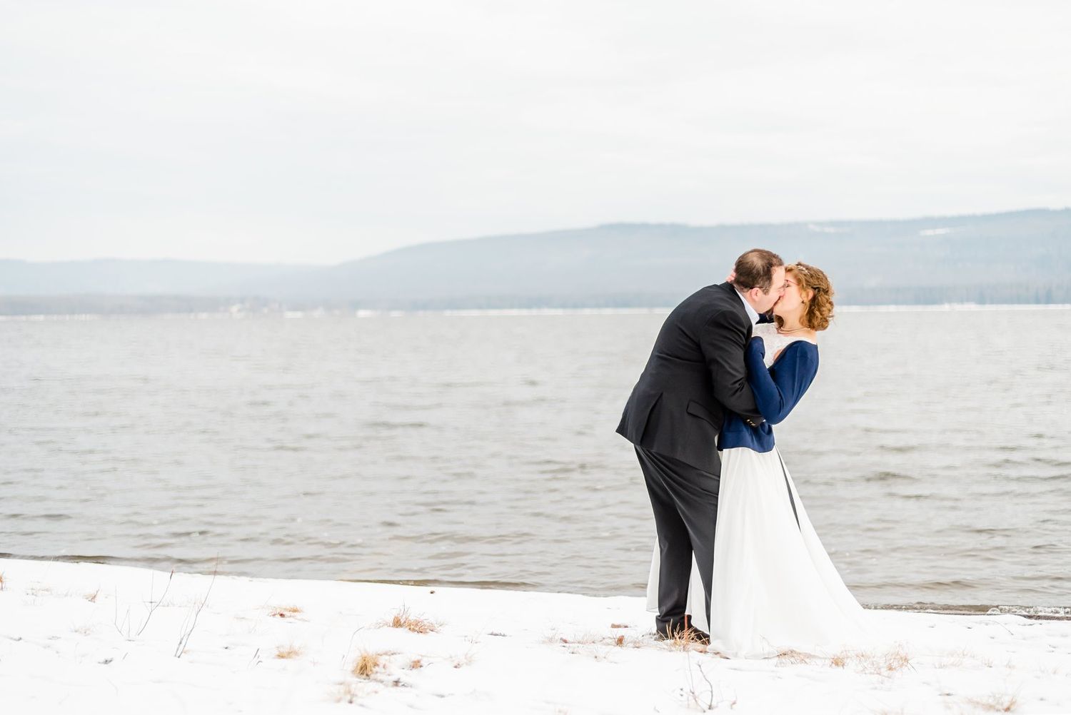 Bride and groom share a romantic kiss on a snowy lakeside shore during a winter wedding photo session.