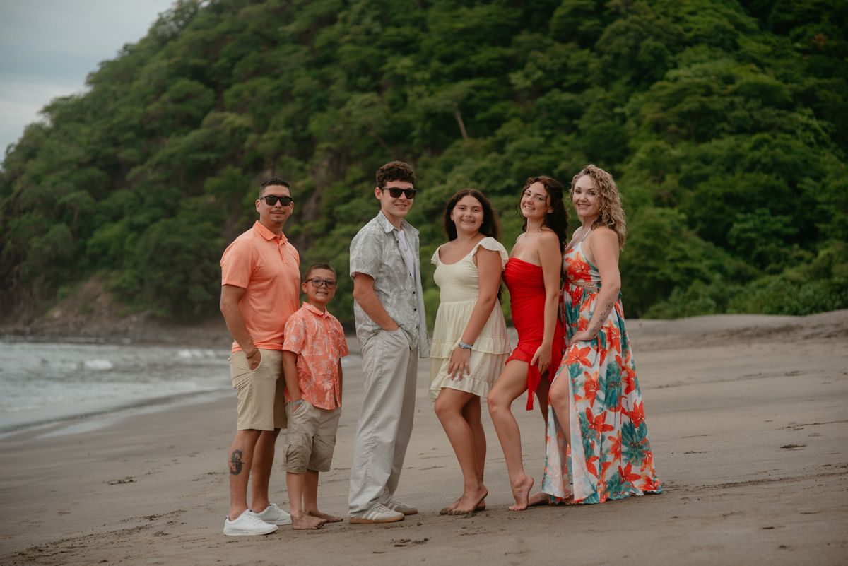 Family of six posing together on a tropical beach with lush green hills in the background.
