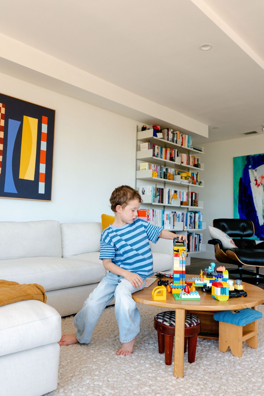 Young boy in striped shirt plays with colorful Lego blocks on a round table in a modern living room.