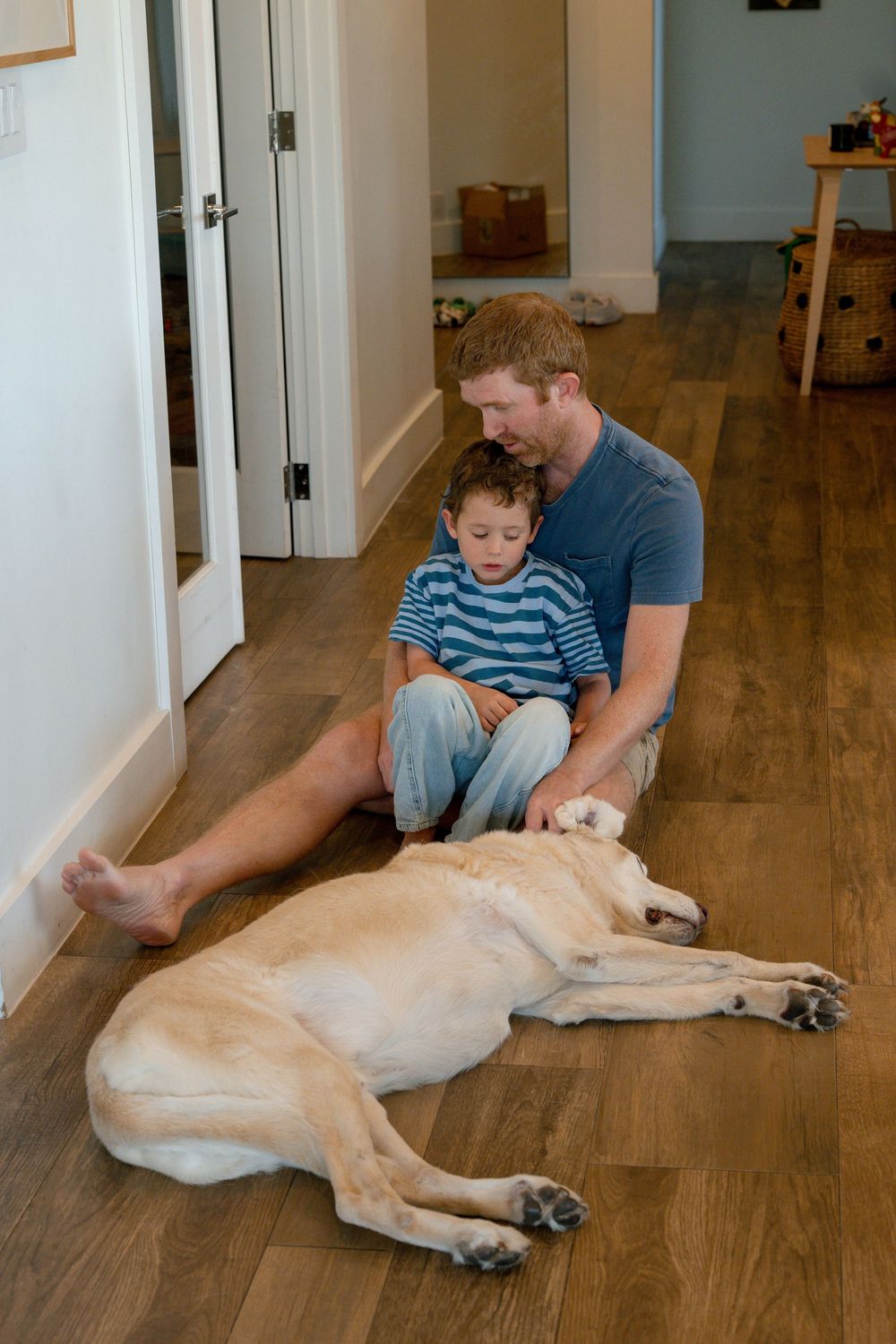 Father and young child sit together on hardwood floor with large yellow Labrador dog lying beside them indoors.