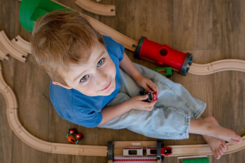 Red-haired boy in blue shirt smiles up at camera while playing with wooden train set on floor.