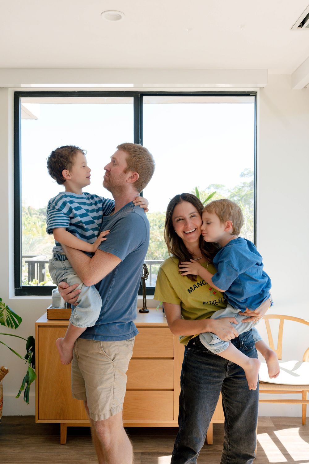 Happy family of four with parents holding two young boys sharing kisses in bright modern home.