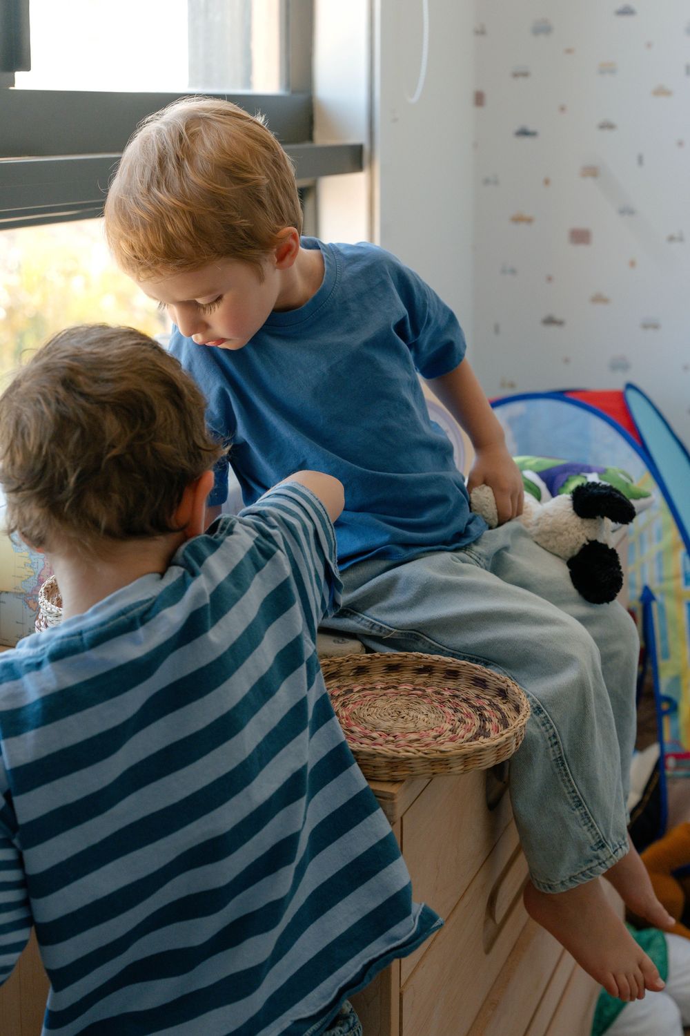Two young boys play together in a colorful kids bedroom, one sitting on a wooden storage unit.