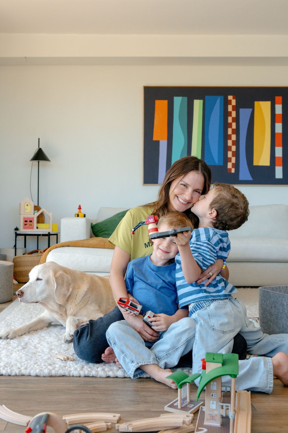 Mother smiling with two young boys and a golden retriever on a rug in a modern living room with colorful wall art.