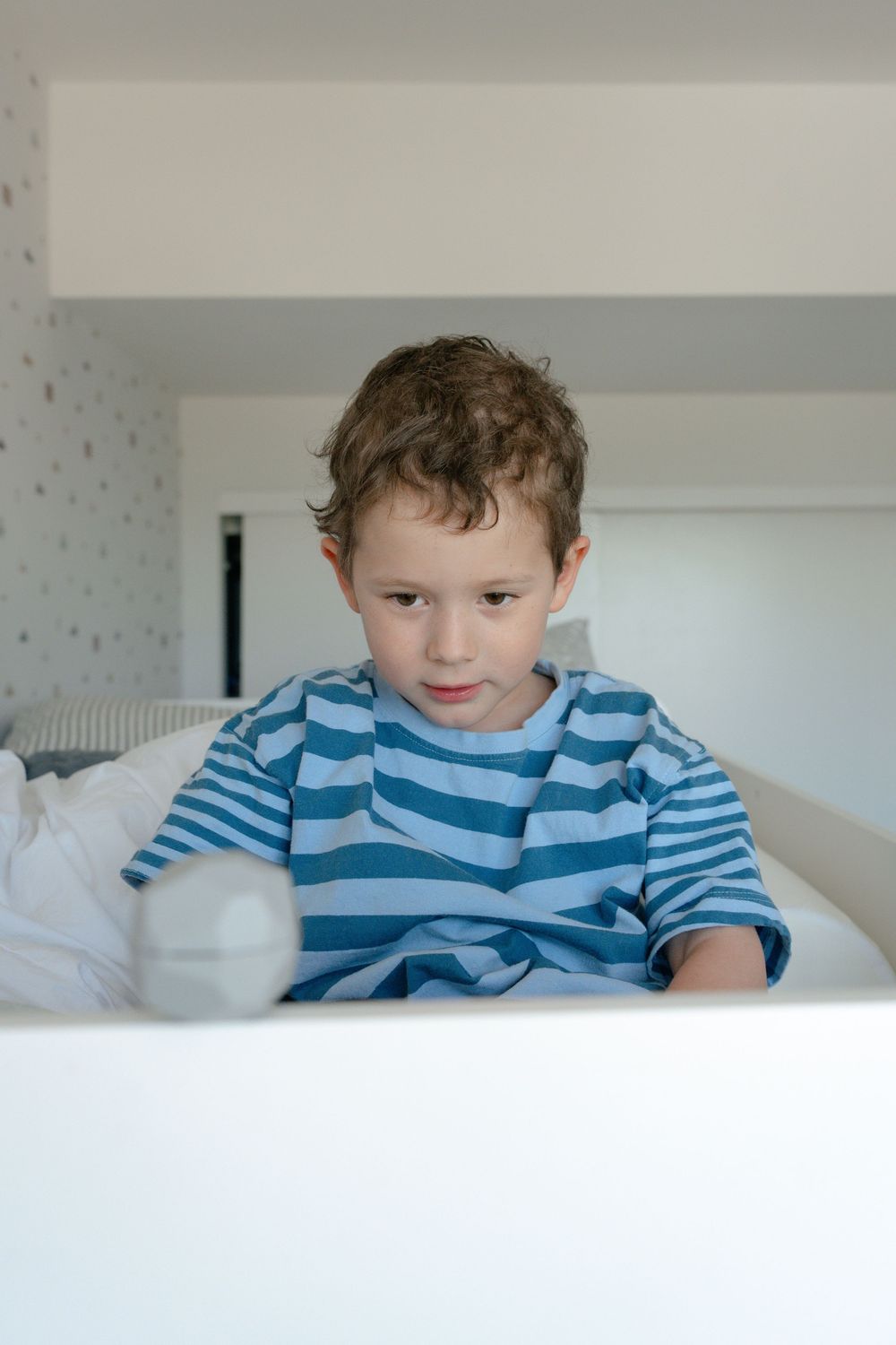 Young curly-haired boy in striped shirt sitting in white bed looking at laptop screen.