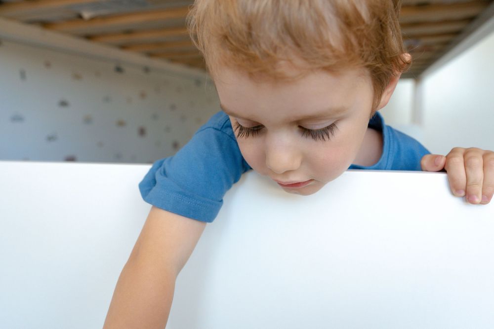 Curious toddler in blue shirt peering over edge of white furniture indoors.