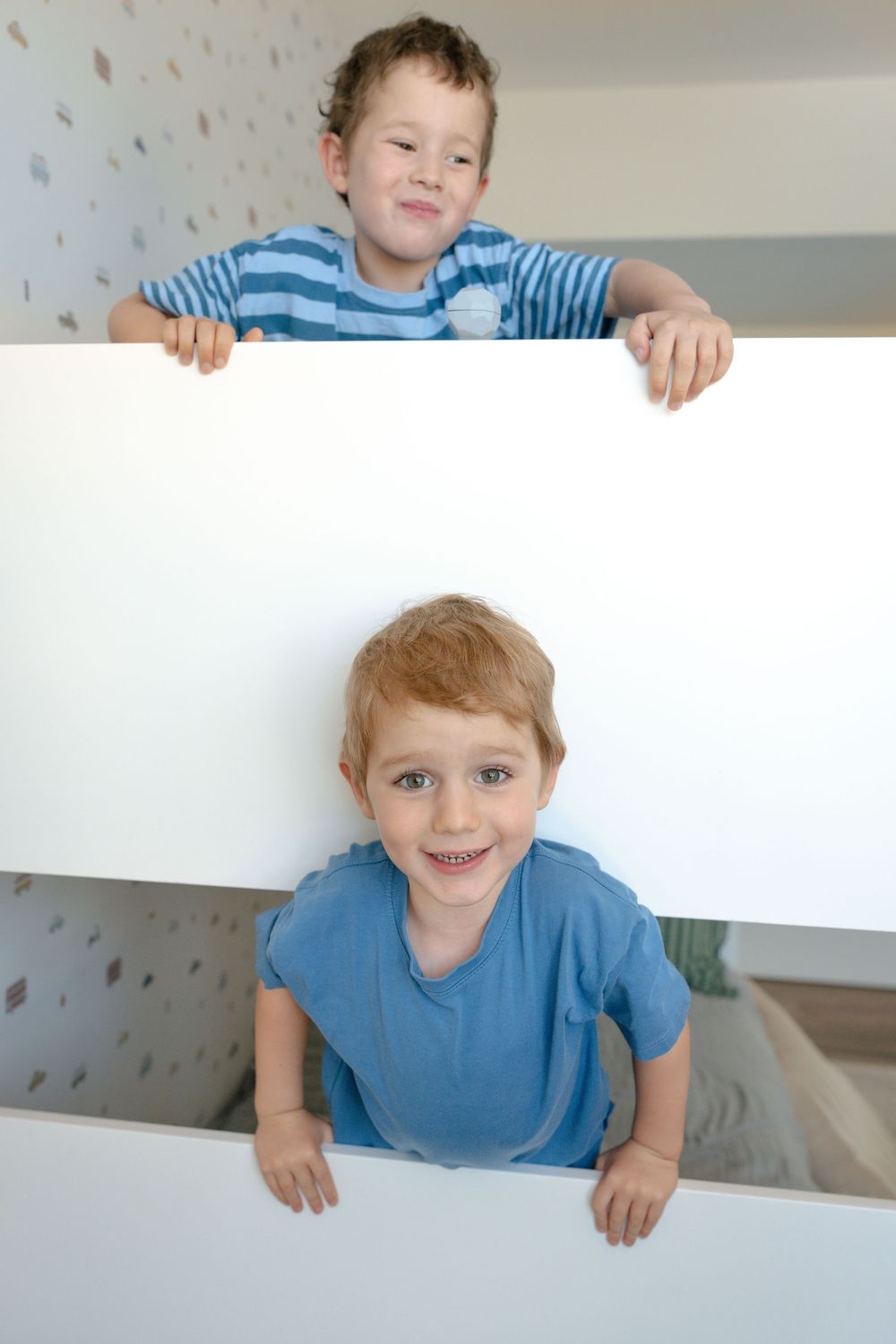 Two young boys in blue shirts peek over a white bunk bed railing, smiling at the camera.