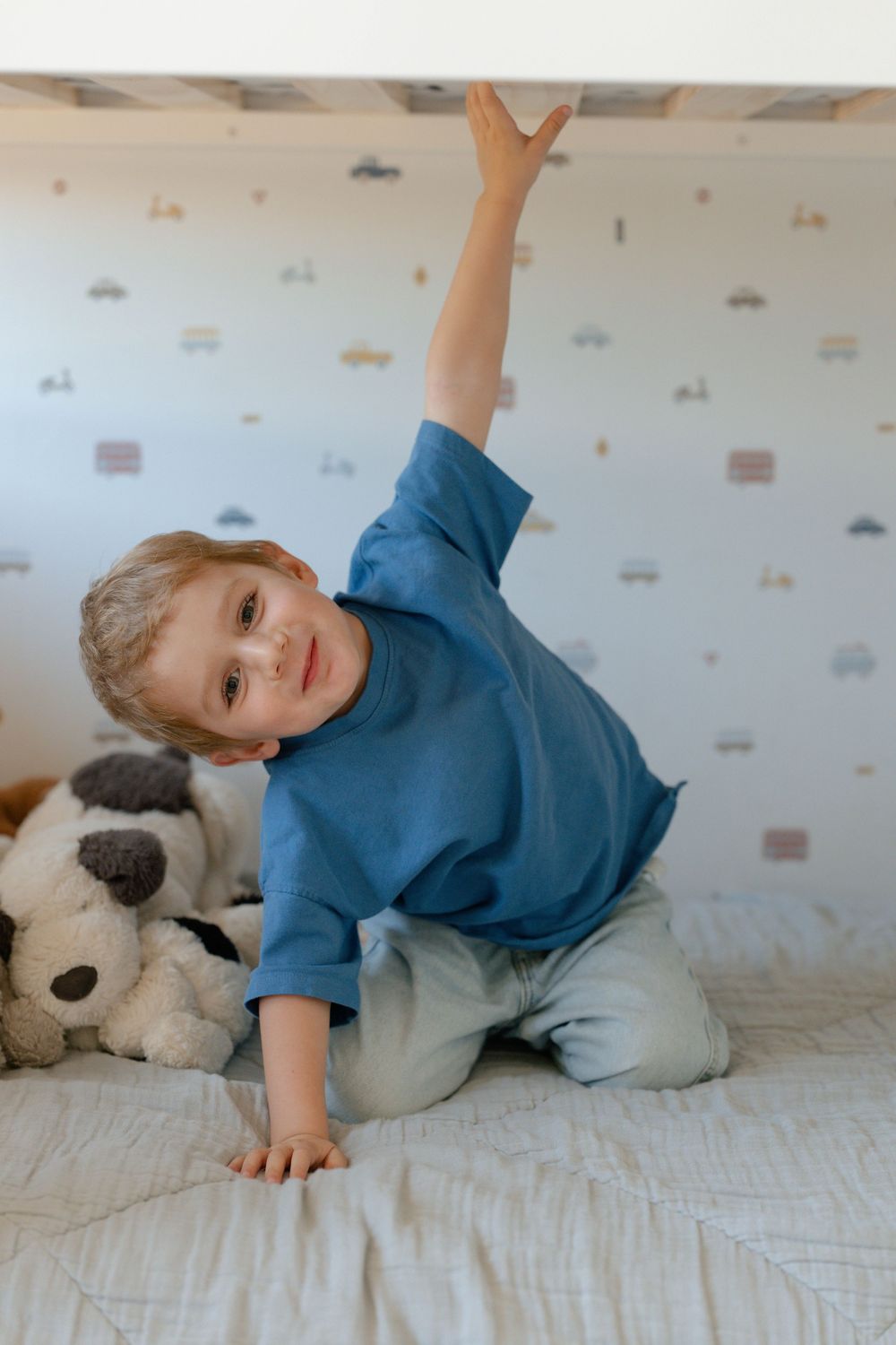 Young boy in blue shirt reaches up while kneeling on a bed with stuffed animals and car wallpaper.