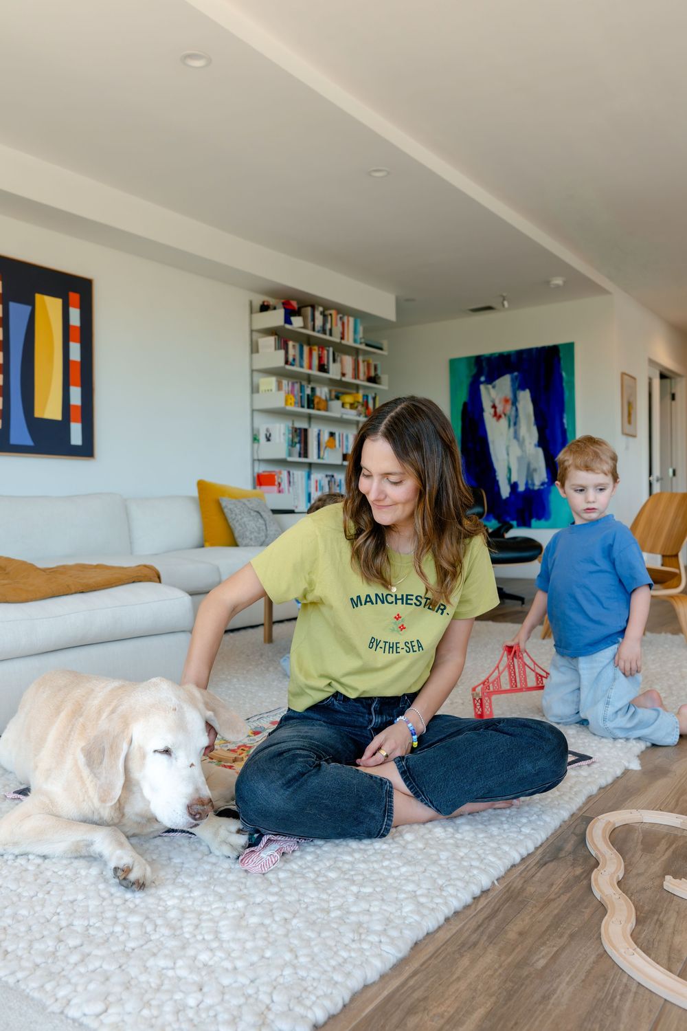 Woman in green t-shirt sitting on rug petting golden retriever while toddler plays with wooden train set nearby.