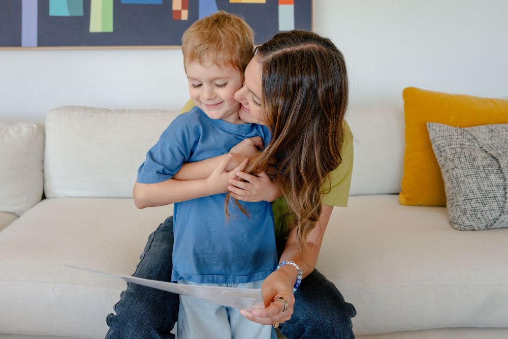 Woman hugs smiling red-haired boy in blue shirt on a white couch in a bright living room.