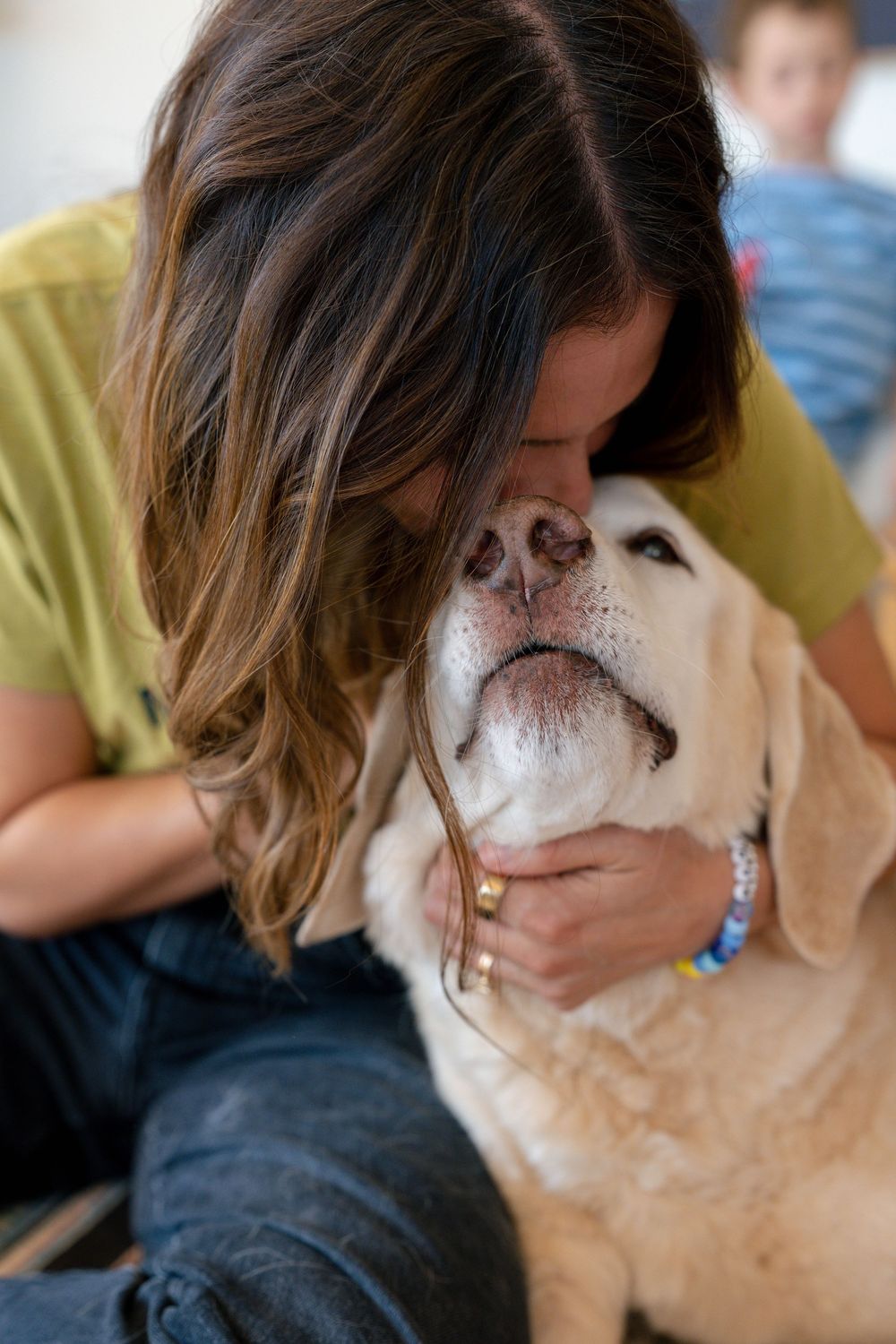 Young woman with brown hair affectionately nuzzling a golden Labrador dog while wearing a green shirt.