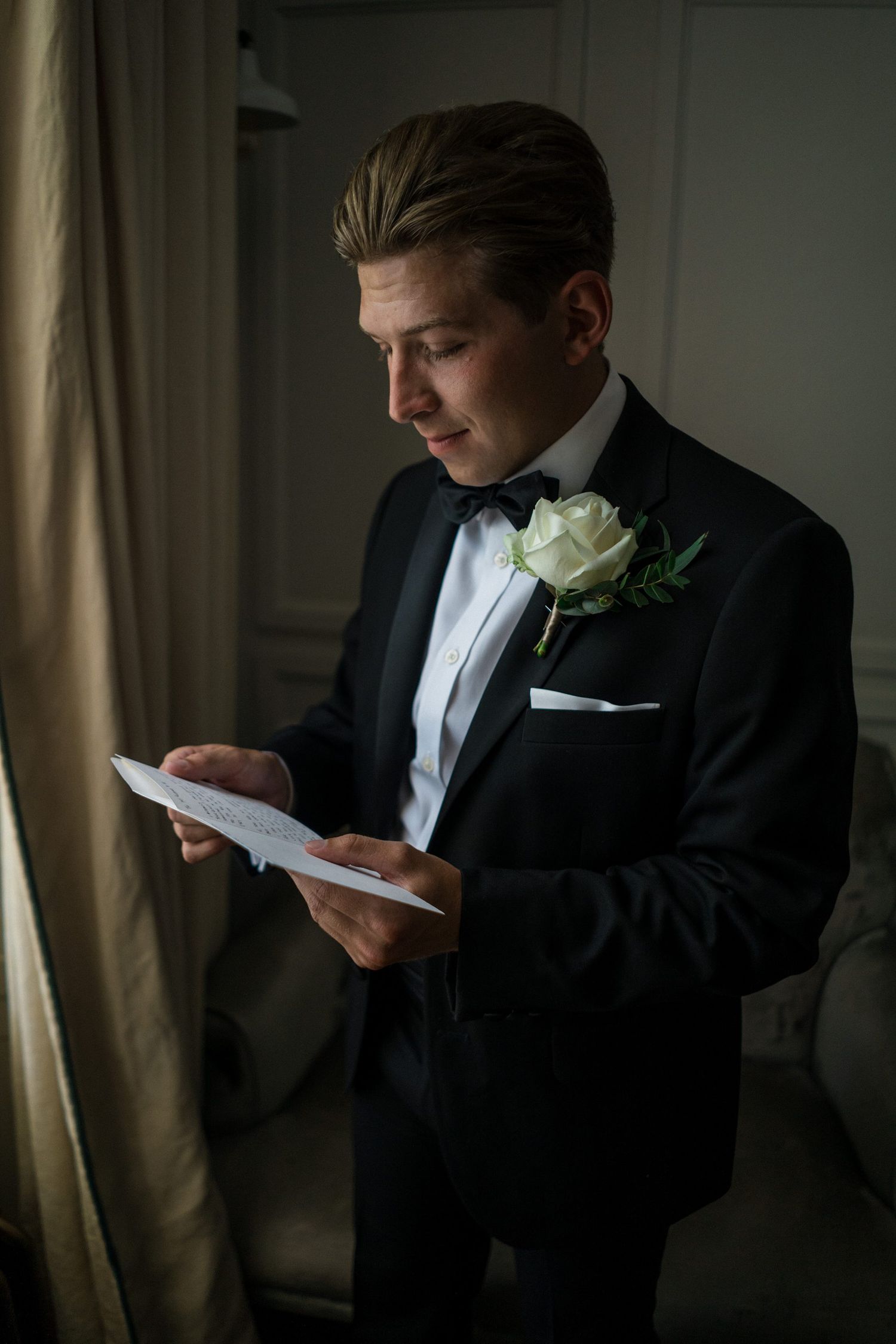 Groom in black tuxedo with white rose boutonniere reading a letter near a window on his wedding day.