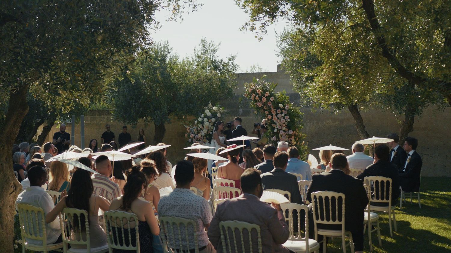 Outdoor wedding ceremony with guests seated on white chairs under olive trees, featuring a floral arch altar.
