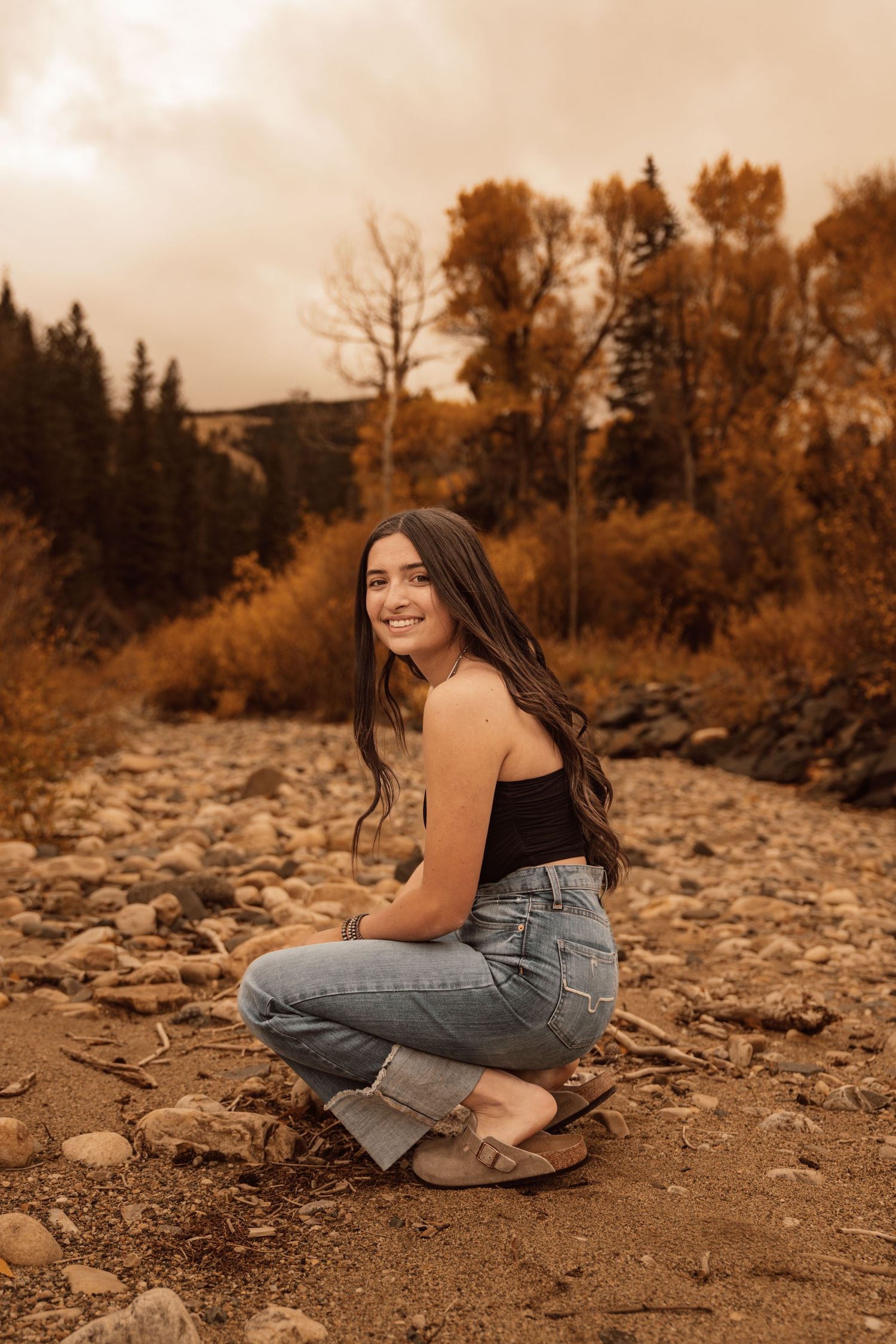 Smiling redhead woman in yellow ruffled dress on autumn dirt path surrounded by golden trees.