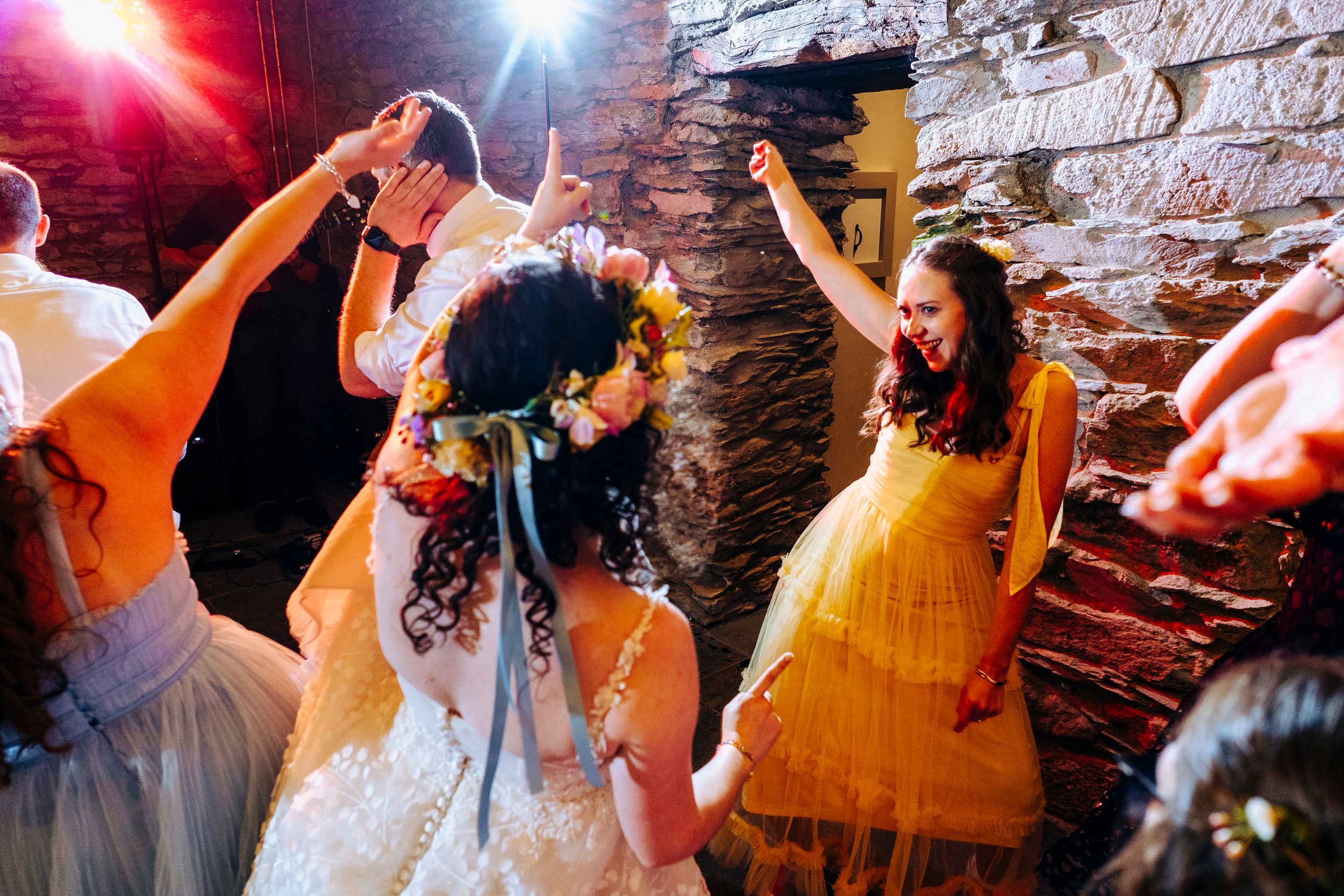 A joyful bride wearing a floral crown dances with guests in a vibrant, warmly lit rustic wedding venue.