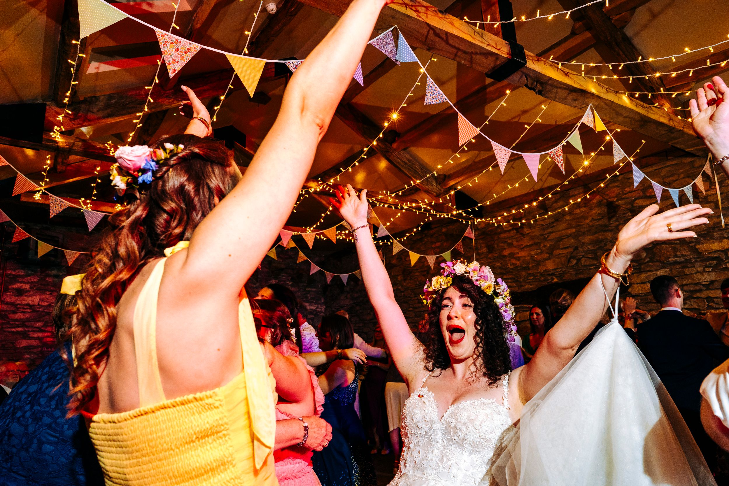 Bride in floral crown dancing joyfully with bridesmaid in yellow dress at colorful wedding reception with bunting.