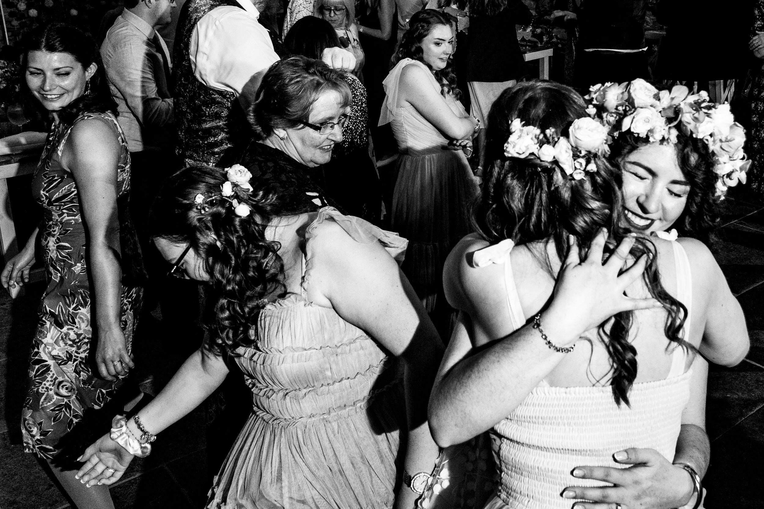 Black and white photo of wedding guests hugging and dancing joyfully, including a bride wearing a floral crown.